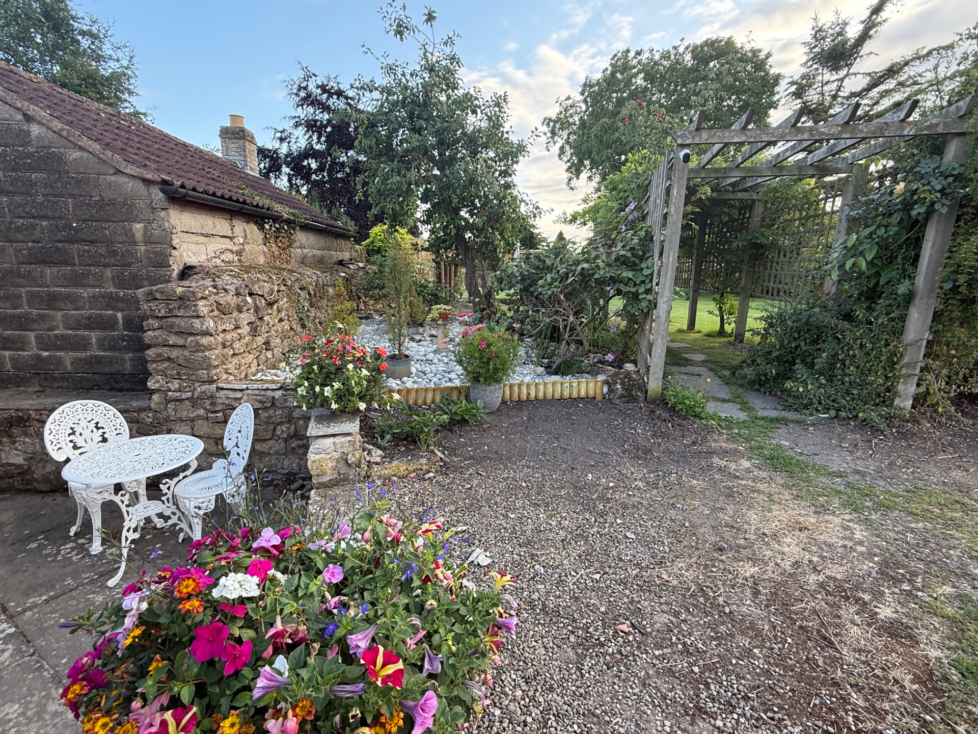 Cottage garden with stone walls, a white cafe table and flowering pots beside a wooden pergola