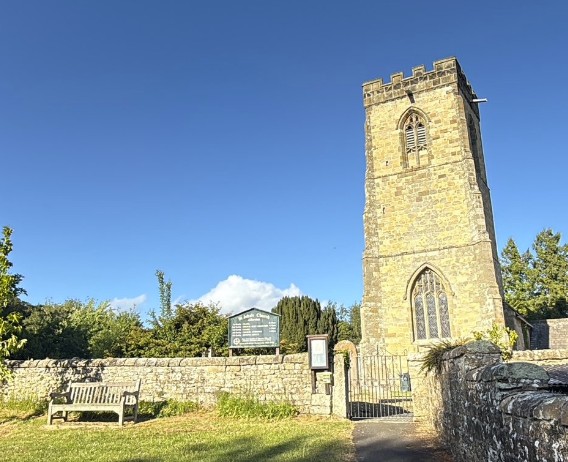 The battlemented stone tower of St John's Church, Allerston, above the churchyard wall and village bench on a clear summer day