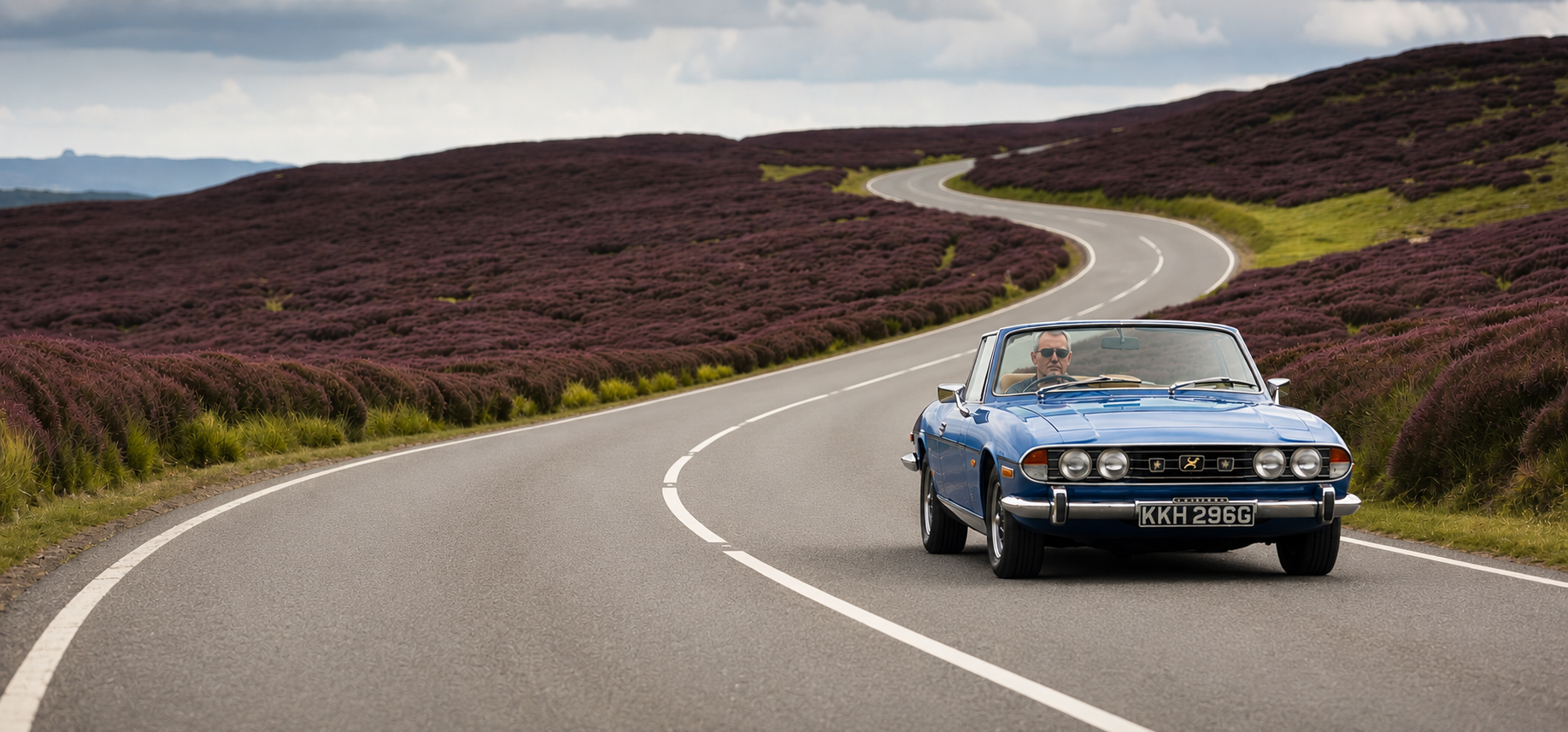 A blue Triumph Stag convertible on a winding road through purple heather moorland in the North York Moors