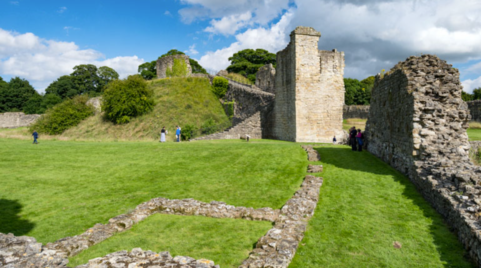 The ruined stone walls and motte of Pickering Castle on a bright day, with visitors exploring the grassy bailey