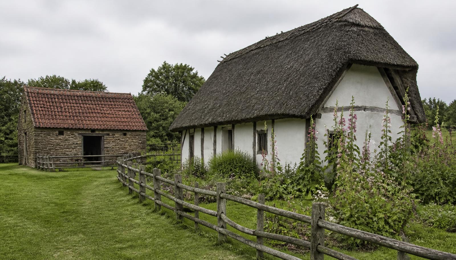 A thatched cruck-framed cottage with a cottage garden and a pantile-roofed stone workshop on the grass of an open-air folk museum in Yorkshire