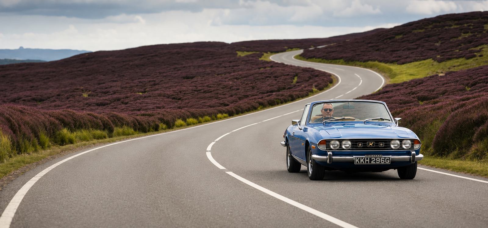 A blue Triumph Stag convertible on a winding moor road through purple heather in the North York Moors