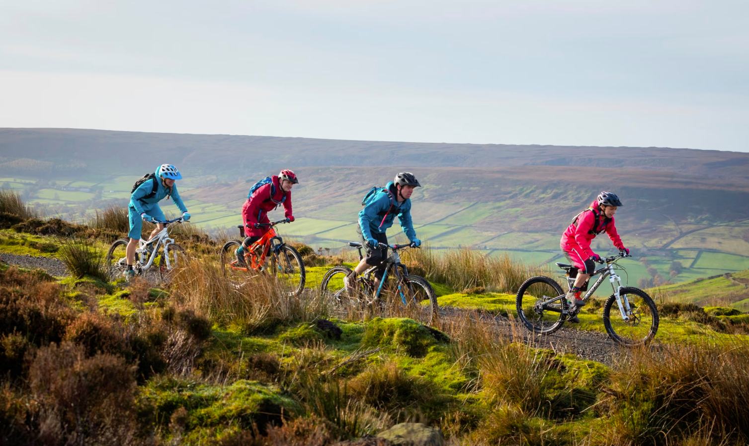Four mountain bikers in colourful jackets riding a singletrack trail across moorland above a green valley in the North York Moors