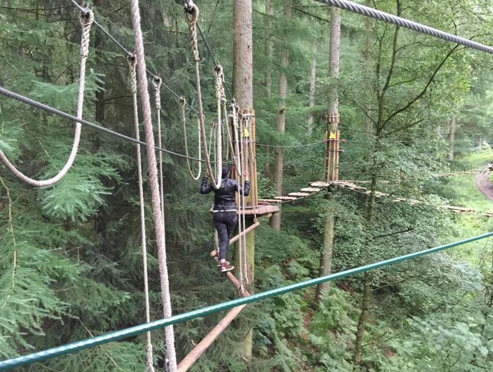 A visitor in a harness crossing a wobbly rope walkway on the Go Ape high-ropes course at Dalby Forest, with tall conifers behind
